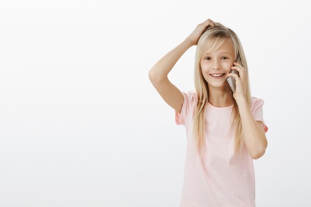 Meisje heeft geen idee, weet niet hoe ze moet antwoorden. Verward schattige jonge dochter in roze t-shirt, hoofd krabben en vrolijk glimlachend tijdens het praten op smartphone, ondervraagd en onbewust