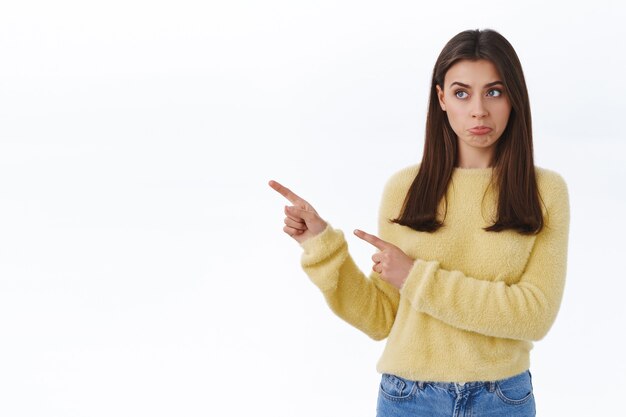 Meisje heeft spijt dat ze een goede kans mist, somber pruilend, kijkend en wijzend met de vingers naar links met jaloerse overstuur of teleurgestelde emoties op het gezicht, staande witte muur mokkend