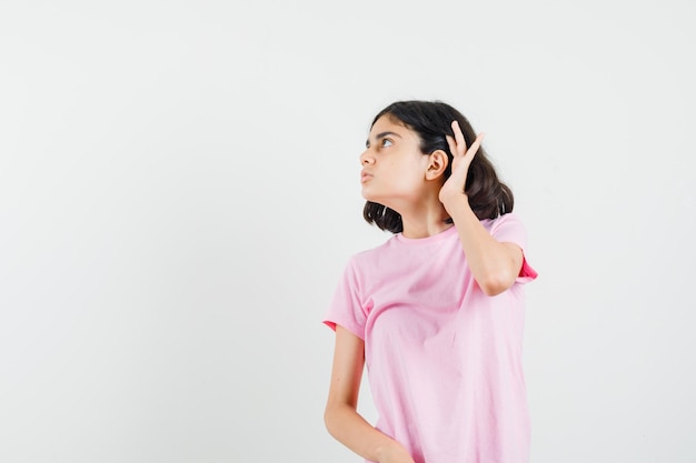 Meisje houdt hand achter oor in roze t-shirt en kijkt nieuwsgierig, vooraanzicht.