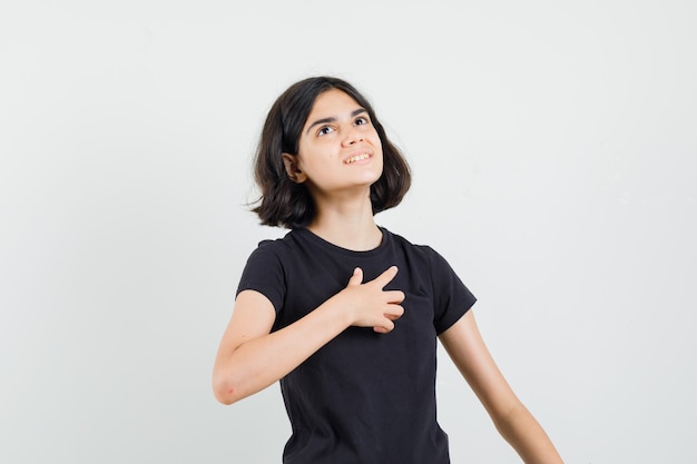 Meisje houdt hand op de borst in zwart t-shirt en kijkt hoopvol. vooraanzicht.