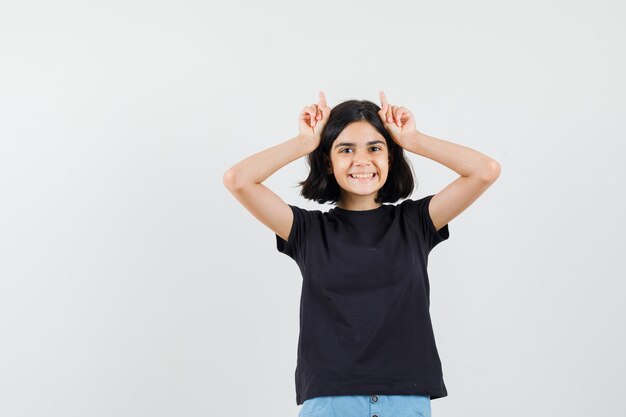 Meisje houdt vingers boven het hoofd als hoorns van de stier in zwart t-shirt, korte broek en op zoek grappig, vooraanzicht.