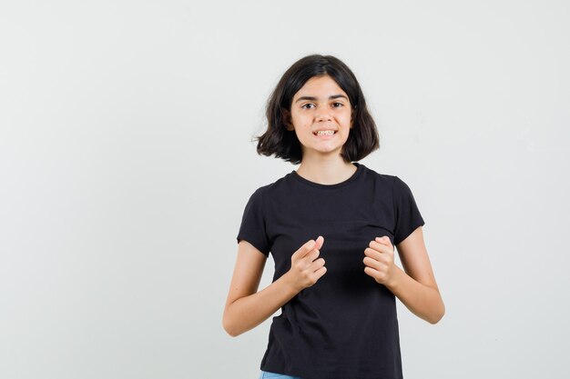 Meisje houdt vuisten op de borst in zwart t-shirt en kijkt vrolijk, vooraanzicht.