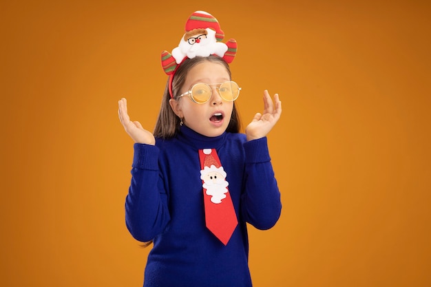 Meisje in blauwe coltrui met rode stropdas en grappige kerstrand op hoofd verward en verrast met opgeheven armen