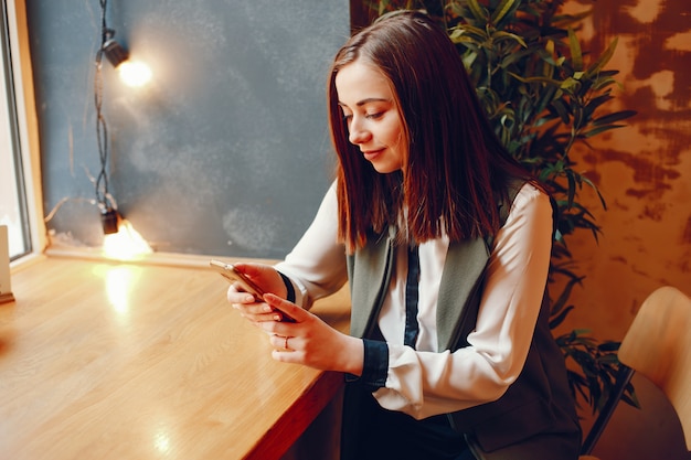 meisje in een witte blouse zitten aan de tafel in de buurt van het venster in het café en houdt de telefoon