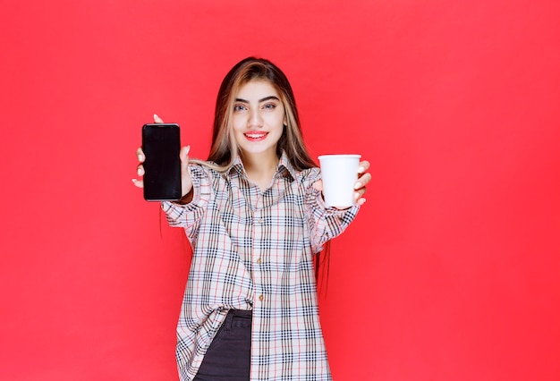 Meisje in geruite trui die een kopje drank vasthoudt en haar smartphone laat zien