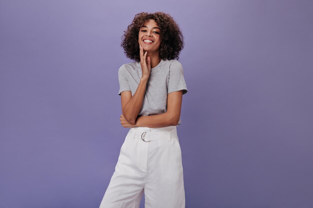 Meisje in grijze tshirt lacht op geïsoleerde achtergrond Vrolijke krullende vrouw in witte korte broek lachen op paarse achtergrond