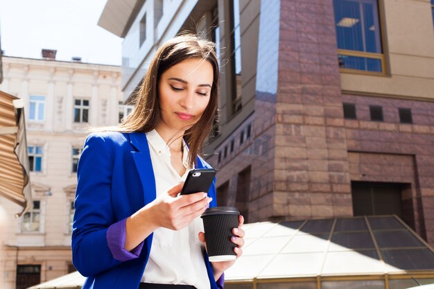 Meisje in helderblauwe jas staat met smartphone en koffie op de straat