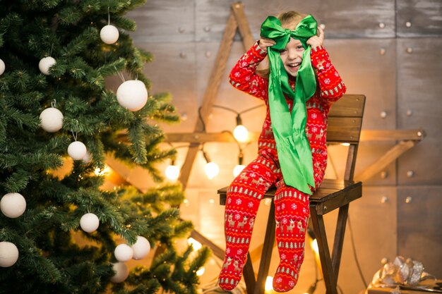 Meisje in pyjama&#39;s door de kerstboom op een houten stoel