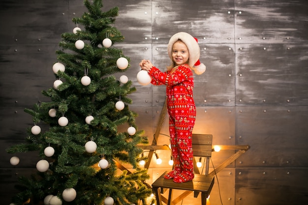 Meisje in pyjama's door de kerstboom op een houten stoel