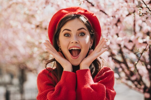 Meisje in rode baret in vreugdevolle schok kijkt naar camera tegen de achtergrond van sakura. Verrast groenogige vrouw in trui poseren in bloeiende tuin