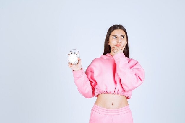 Meisje in roze pyjama met een wekker en ziet er bedachtzaam en verward uit.