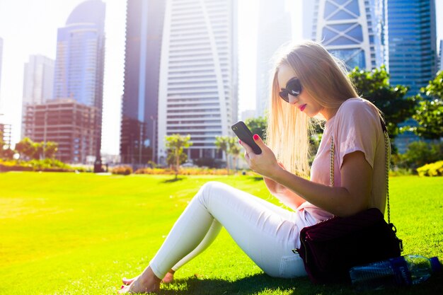 Meisje in roze shirt en witte jeans controleert haar telefoon op het gras in het park zitten