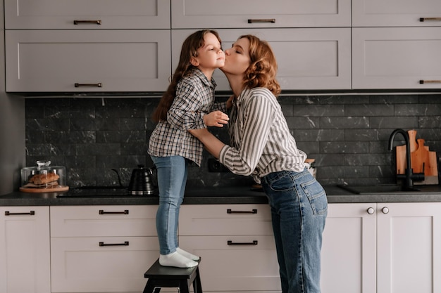 Meisje in spijkerbroek en shirt staat op stoel in de keuken en moeder kust haar op de wang