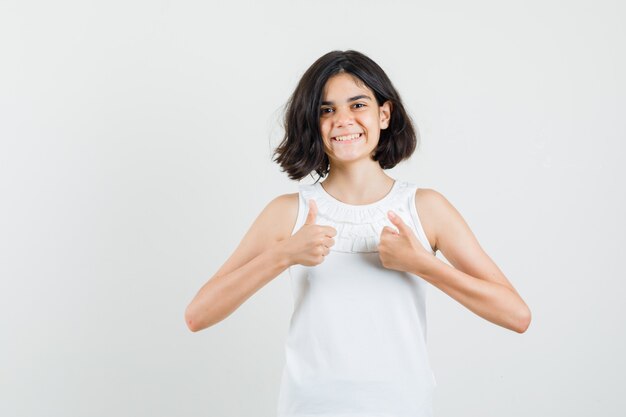 Meisje in witte blouse die dubbele duimen toont en vrolijk, vooraanzicht kijkt.