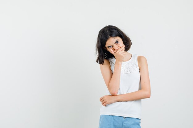 Meisje in witte blouse, korte broek die zich in het denken stelt en ernstig, vooraanzicht kijkt.