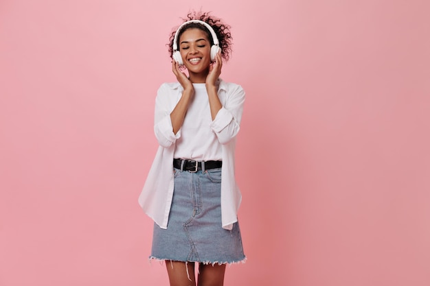 Meisje in witte t-shirt en denim rok geniet van muziek op roze achtergrond gelukkig donkerhuidige vrouw in tee glimlachend op geïsoleerde achtergrond