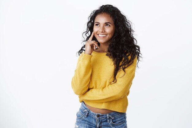 Meisje kijkt tevreden en knikt met acceptatie. Knappe vrouw met een donkere huidskleur met krullend haar, wang aanraken en nadenkend glimlachen, kijk tevreden linkerbovenhoek, zoals kopieerruimte