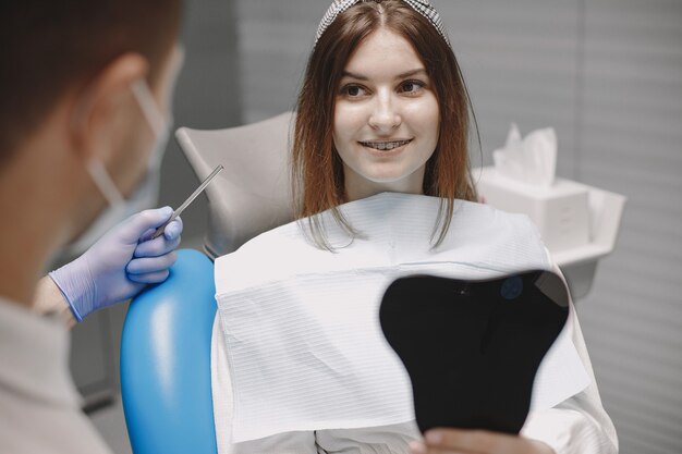 Meisje met beugels die in de spiegel naar zichzelf kijken op het kantoor van de tandarts. Stomatoloog met blauwe handschoenen en gezichtsmasker