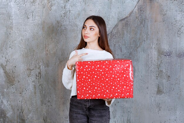 Meisje met een rode geschenkdoos met witte stippen erop en ziet er attent en dromerig uit.