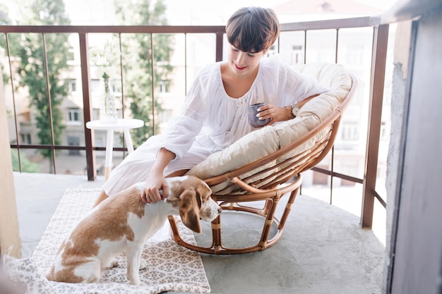 Meisje met elegante witte manicure beagle hond aaien terwijl u geniet van koffie in de ochtend. Prachtige jonge dame in zomerjurk met theetijd op terras en spelen met puppy.