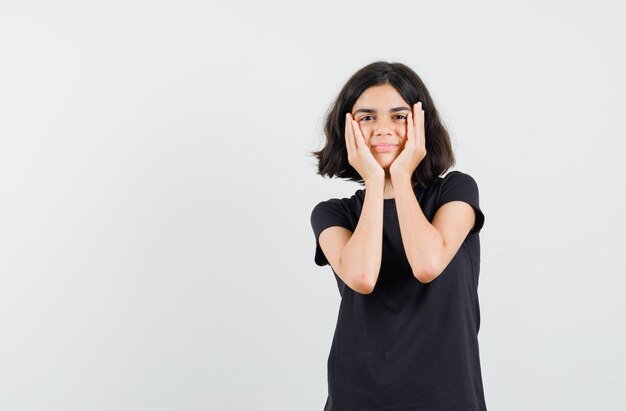 Meisje met handpalmen op de wangen in zwart t-shirt en ziet er mooi uit, vooraanzicht.