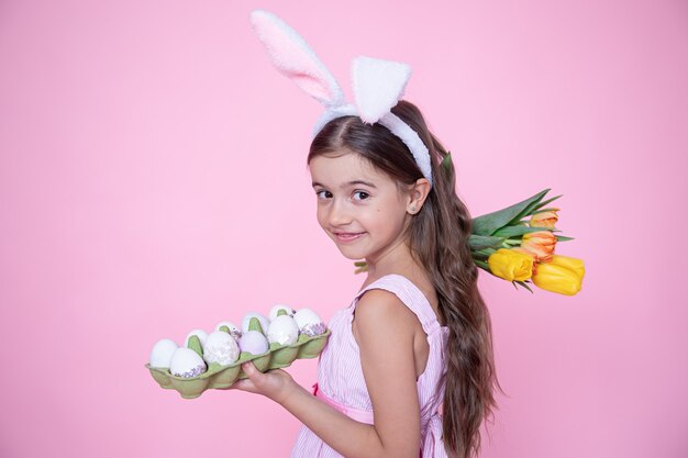 Meisje met paashaasoren houdt een boeket tulpen en een dienblad met eieren in haar handen op een roze muur.