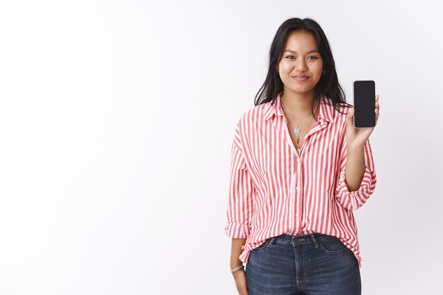 Meisje met smartphone stelt ze voor om te kopen. Opgetogen gelukkige aantrekkelijke jonge Polynesische vrouw in gestreepte roze blouse met mobiele telefoon presenteren app op gadget scherm op witte achtergrond