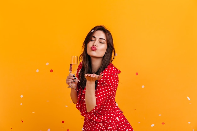 Meisje met veel gevoel voor humor heeft plezier op feestje met een glas champagne. Vrouw in lichte jurk stuurt kus tegen muur van confetti.