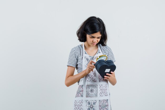 Meisje met vergrootglas over geschenkdoos in t-shirt, schort