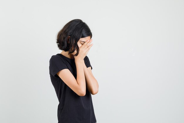 Meisje omklemde handen op gezicht in zwart t-shirt en kijkt verdrietig. vooraanzicht.