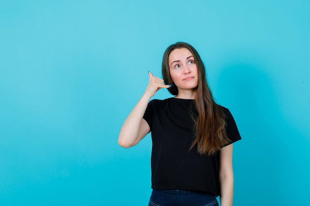 Meisje opzoeken toont telefoongebaar door hand in de buurt van oor op blauwe achtergrond te houden