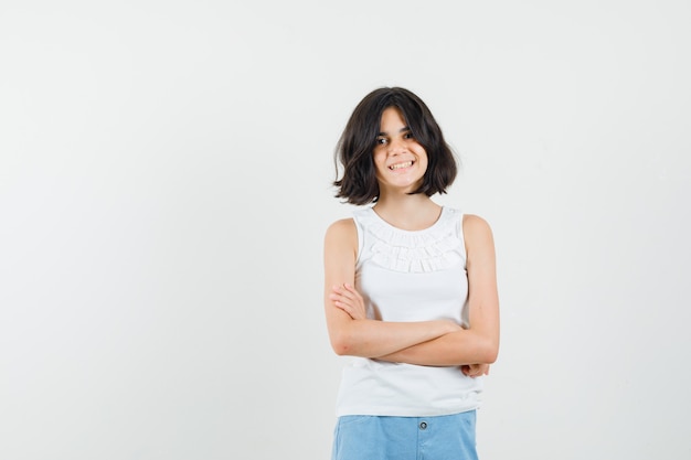 Meisje permanent met gekruiste armen in witte blouse, korte broek en op zoek vrolijk. vooraanzicht.