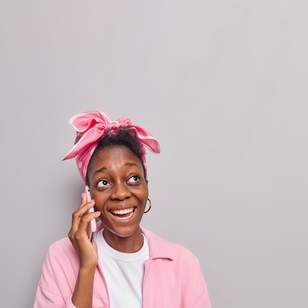 meisje praat op mobiele telefoon heeft positieve uitdrukking beantwoordt telefoontje beste vriend via mobiel draagt roze jas hoofddoek vastgebonden op hoofd staat binnen