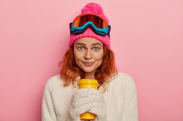 Meisje snowboarder draagt warme winter outfit, witte wanten, houdt afhaalkoffie, geïsoleerd op roze achtergrond