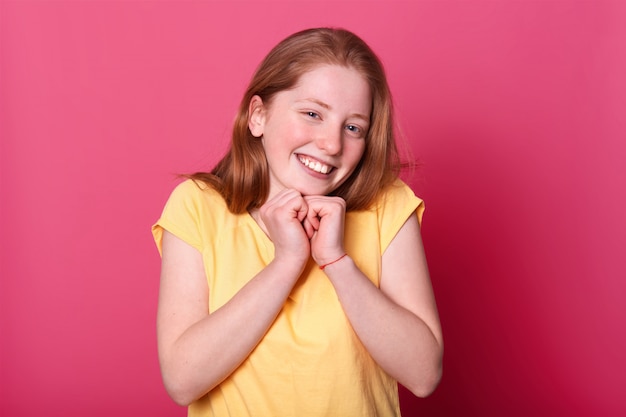 meisje staat op roze muur, gekleed casual gele t-shirt, heeft lange rechte haren, houdt beide handen onder de kin