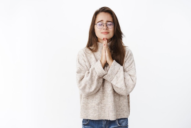 Meisje voelt zich bezorgd en onzeker fronsend en emotioneel als gesloten ogen hand in hand in gebed, wensen biddend trouw met hoop dat dingen beter worden, nerveus en ongelukkig op een grijze muur staan.