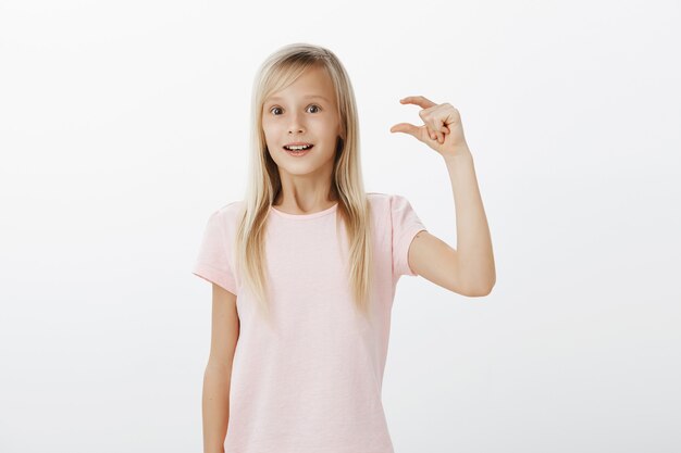 Meisje voelt zich vrolijk en opgewonden, deelt indrukken na een bezoek aan de dierentuin. Schattige blonde dochter in roze t-shirt, hand opsteken en vormgeven van klein of klein ding met bewonderende uitdrukking over grijze muur