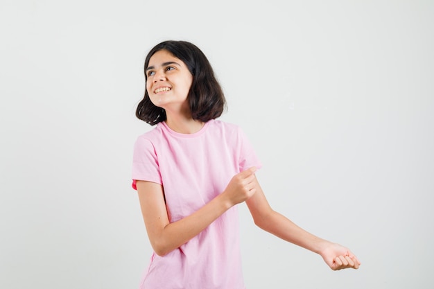 Meisje wijst opzij, kijkt omhoog in roze t-shirt en kijkt vrolijk. vooraanzicht.