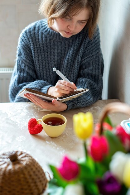 Meisje writinig in een notitieboekje, gebreid rood hart en een kopje thee op tafel. Keuken