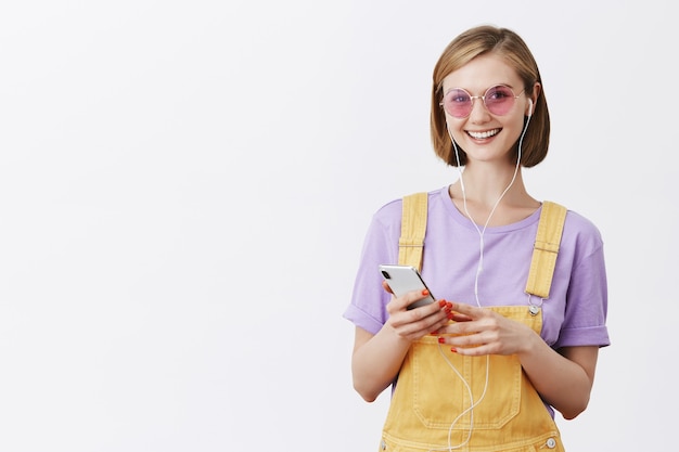 Meisje zetten oortelefoons permanent in gele tuinbroek en zonnebril, smartphone te houden en glimlachen