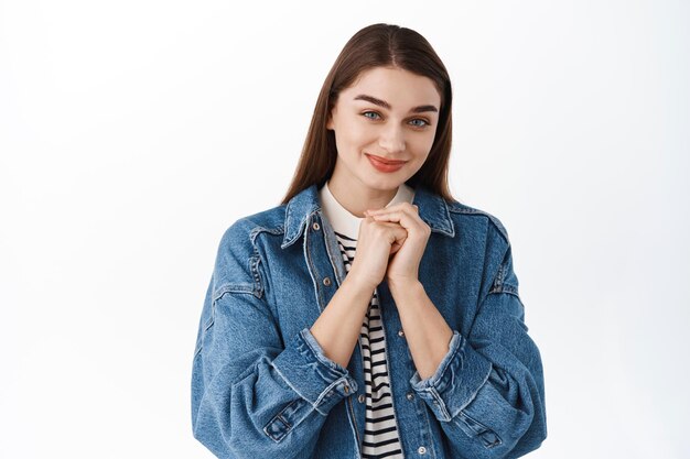 Meisje ziet er terughoudend en schattig uit, maakt een dom romantisch gezicht, houdt handen dicht bij het hart en glimlacht onschuldig naar de camera, engel kijkt, staande op een witte achtergrond