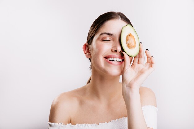 Meisje zonder make-up houdt een halve avocado vast en bedekt een deel van het gezicht. Vrij vrouwelijk model met donker haar leidt een gezonde levensstijl.