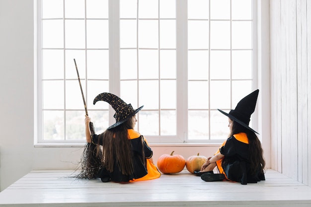 Gratis foto meisjes in halloween-kostuums die met bezem elkaar dichtbij venster bekijken