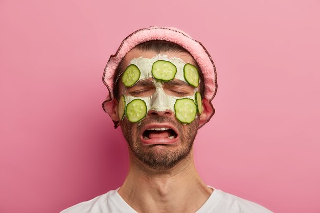Melancholisch mannelijk model heeft zelfgemaakt masker met komkommers op gezicht, moe van het bezoeken van spa-salon, draagt een wit t-shirt en badmuts, kan zich niet ontspannen voelen