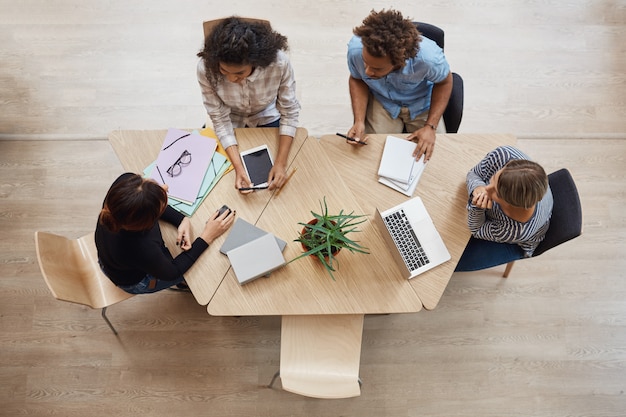 Mening van hierboven van groeps jonge professionele ondernemers die bij lijst in coworking-ruimte zitten, die winsten van laatste teamproject bespreken, gebruikend laptop, digitale tablet en smartphone.