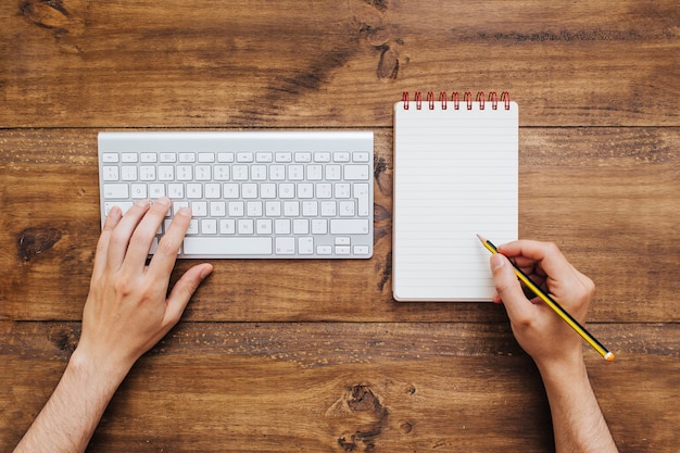 Mens die aan toetsenbord werkt en op blocnote schrijft.