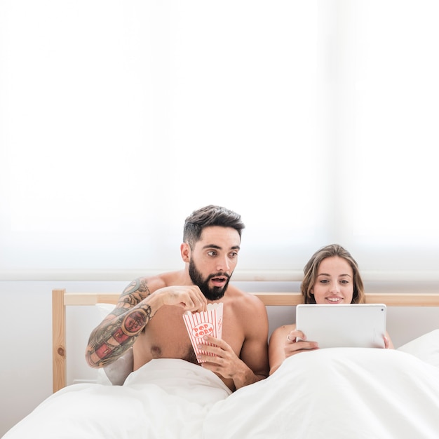 Mens die met popcorn zijn vrouw bekijkt die digitale tablet op bed gebruikt