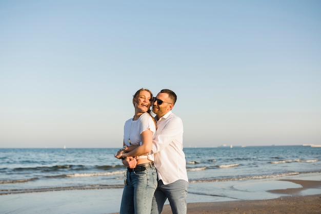 Mens die zijn meisje van achter omhelst status dichtbij het overzees tegen blauwe hemel bij strand