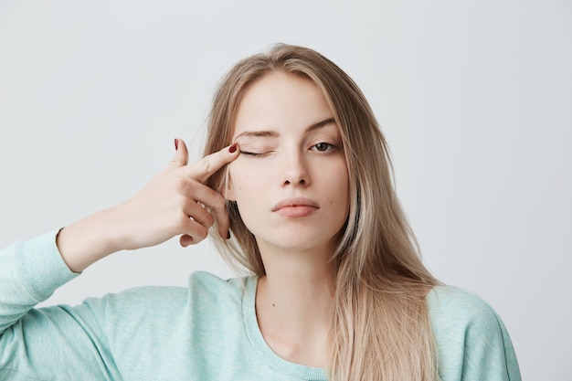 Gratis foto menselijk gezicht uitdrukkingen. doordachte jonge mooie vrouw met blond geverfd steil haar in lichtblauwe trui met lange mouwen aanraken met vingeroog