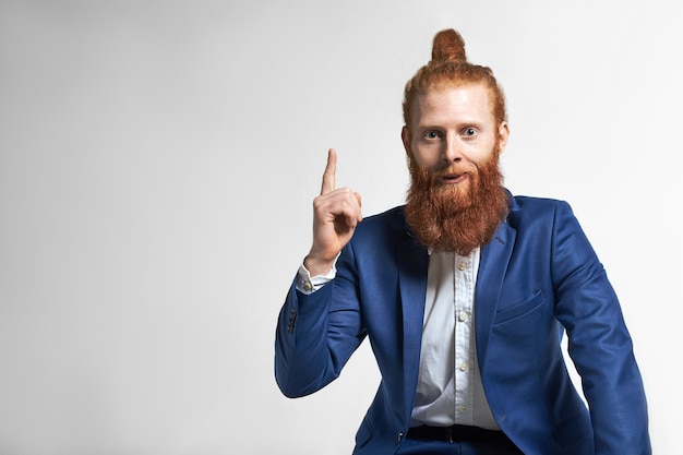 Menselijke emoties, gevoelens en reacties. Portret van knappe emotionele jonge blanke mannelijke werknemer met wazige baard en haarknoop met verrassende blik, wijsvinger wijzend naar de muur van de kopie ruimte